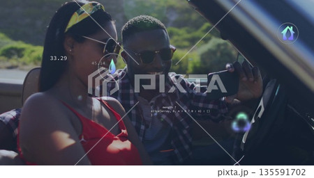 Smiling couple wearing sunglasses capturing selfie in beige convertible on rural road, smartphone 135591702