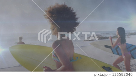 Walking three women wearing bikinis carrying surfboards along sandy beach at sunset, ocean waves 135592040