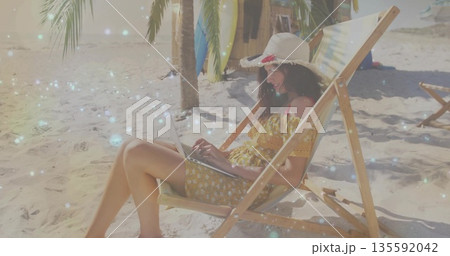 Freelancer wearing straw sun hat and yellow dress working on laptop on sandy beach, with surfboards 135592042