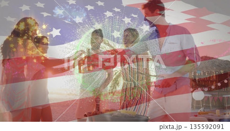 Toasting five friends holding beer bottles on beach at sunset, with tiki hut, grill, flag overlay 135592091