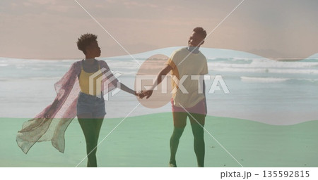 Walking couple holding hands along shoreline near ocean, with sunglasses and pink duster fluttering 135592815