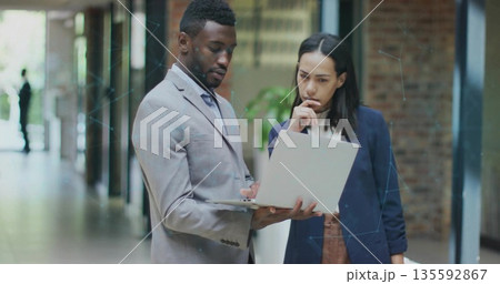 Reviewing colleagues in suit and blazer working on silver laptop in office corridor, with plant Reviewing colleagues in suit and blazer working on silver laptop in office corridor, with plant 135592867
