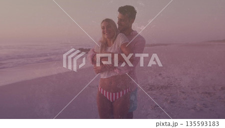 Embracing couple wearing swimwear gazing at horizon on sandy shoreline by sea at dusk, pink sky Embracing couple wearing swimwear gazing at horizon on sandy shoreline by sea at dusk, pink sky 135593183