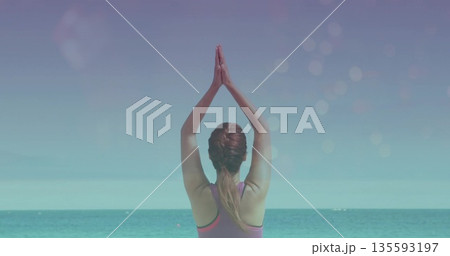 Woman in purple-pink top practicing yoga prayer pose on beach shore facing turquoise sea with bokeh Woman in purple-pink top practicing yoga prayer pose on beach shore facing turquoise sea with bokeh 135593197