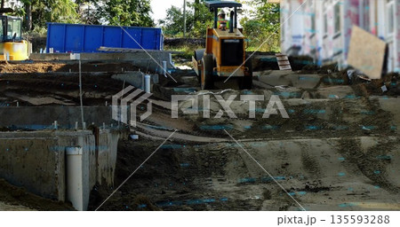Operating road roller compactor smoothing dirt at construction site, with footings, PVC pipes 135593288