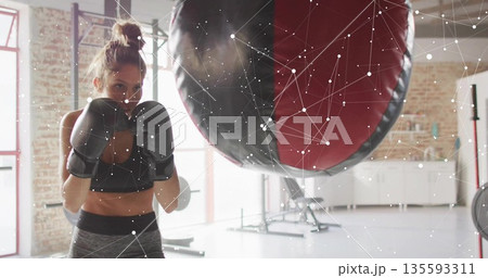 Punching female athlete wearing sportswear striking heavy bag in fitness studio, with boxing gloves 135593311