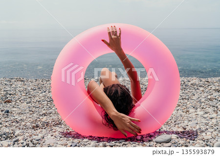 Woman beach vacation lady relaxing inside a large pink inflatable ring on a pebble shore by the calm sea during summer Woman beach vacation lady relaxing inside a large pink inflatable ring on a pebble shore by the calm sea during summer 135593879