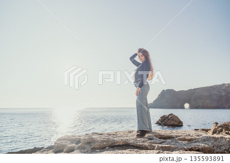 Woman coast sea standing on a rocky shore enjoying the bright morning sun over the calm ocean water 135593891