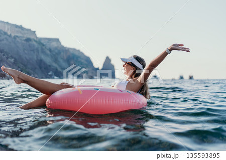Woman, donut, ocean relaxing on a pink float in the sea during summer vacation with scenic cliffs and copy space Woman, donut, ocean relaxing on a pink float in the sea during summer vacation with scenic cliffs and copy space 135593895