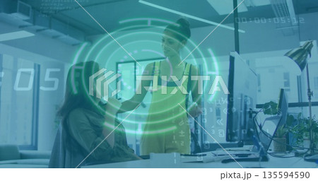 Collaborating woman and coworker visualizing data on monitors in office, with holographic overlay Collaborating woman and coworker visualizing data on monitors in office, with holographic overlay 135594590