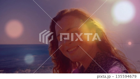 Smiling adult woman standing by sea during golden hour, showing patterned blouse and wavy hair Smiling adult woman standing by sea during golden hour, showing patterned blouse and wavy hair 135594679