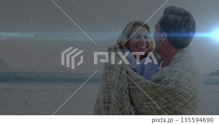 Standing adult couple wrapping in beige patterned blanket on beach, with light scarf and star field 135594690