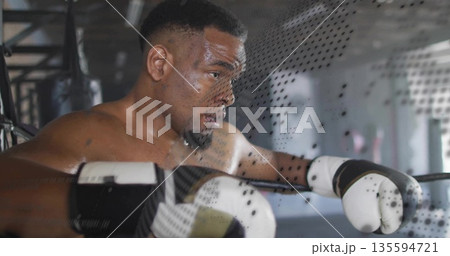 Leaning shirtless male boxer resting arms on boxing ring ropes in gym, with hanging punching bags Leaning shirtless male boxer resting arms on boxing ring ropes in gym, with hanging punching bags 135594721