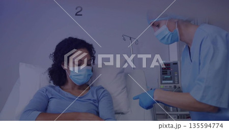 Leaning gloved nurse in blue scrubs pointing at patient arm on hospital bed, with infusion pump 135594774
