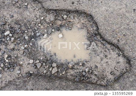 Muddy puddle filling a pothole on a damaged asphalt road surrounded by cracked pavement and loose gravel. Concept of urban infrastructure deterioration Muddy puddle filling a pothole on a damaged asphalt road surrounded by cracked pavement and loose gravel. Concept of urban infrastructure deterioration 135595269