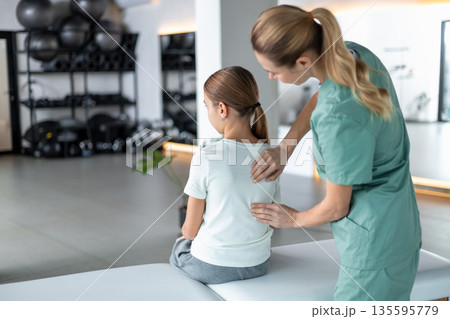 Woman physio in medical uniform checking girl spine in rehab room 135595779