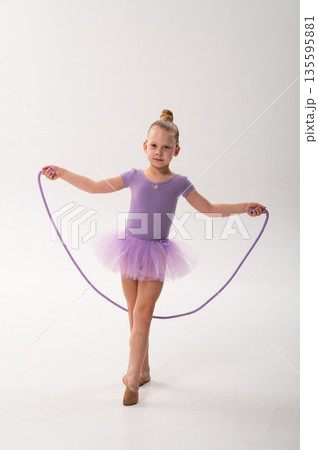 Young ballerina with a cochlear implant stretches a red hoop while posing in a lavender tutu. Inclusive gymnastics, hearing technology, and childhood coordination through rhythmic dance and movement. Young ballerina with a cochlear implant stretches a red hoop while posing in a lavender tutu. Inclusive gymnastics, hearing technology, and childhood coordination through rhythmic dance and movement. 135595881