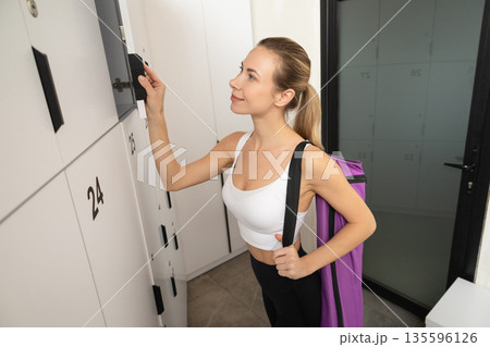 Cute blonde young woman with a violet bag opening locker in gym 135596126