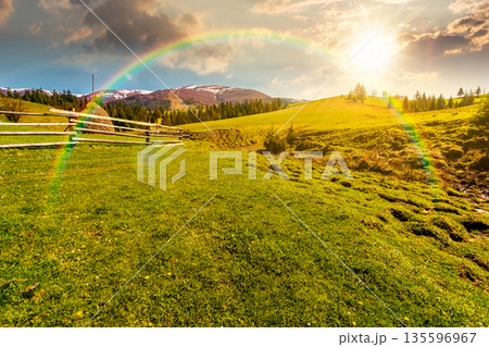 rural landscape in spring at sunset. haystack near wooden fence on green meadow in evening light. spruce forest on rolling hills. mountain with snow tops. timeless nostalgia book cover with rainbow rural landscape in spring at sunset. haystack near wooden fence on green meadow in evening light. spruce forest on rolling hills. mountain with snow tops. timeless nostalgia book cover with rainbow 135596967