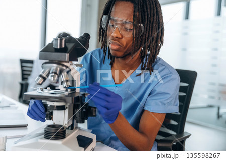 In protective eyewear, with microscope. Black man doctor is in the office In protective eyewear, with microscope. Black man doctor is in the office 135598267