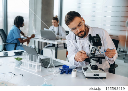 In lab coat, sitting with microscope. Doctors are in the clinic In lab coat, sitting with microscope. Doctors are in the clinic 135598419