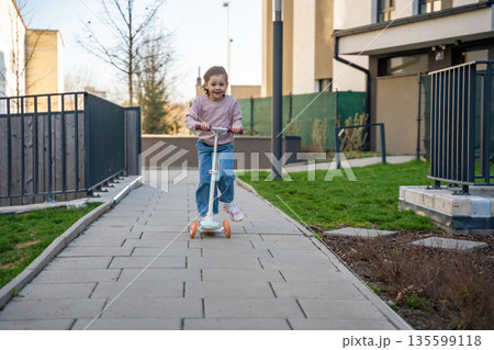 Little girl riding scooter in residential courtyard during spring. Active outdoor play and everyday childhood lifestyle in urban environment. Little girl riding scooter in residential courtyard during spring. Active outdoor play and everyday childhood lifestyle in urban environment. 135599118