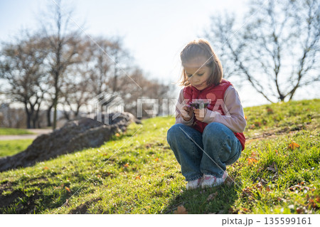 Little girl exploring nature in city park during spring walk. Curious child interacting with soil, leaves and landscape in outdoor environment. 135599161