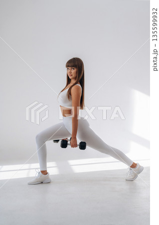 Holding dumbbells and doing squats. Young sportive woman is in the studio 135599932