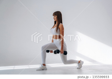 Holding dumbbells and doing squats. Young sportive woman is in the studio 135599933