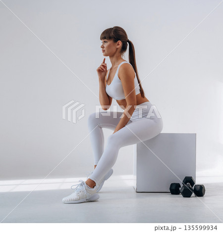 Sitting on the white cube, dumbbells are on the floor. Young sportive woman is in the studio 135599934