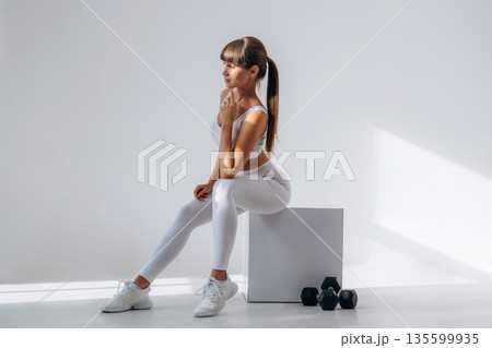 Sitting on the white cube, dumbbells are on the floor. Young sportive woman is in the studio 135599935