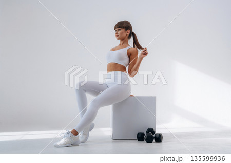 Sitting on the white cube, dumbbells are on the floor. Young sportive woman is in the studio 135599936