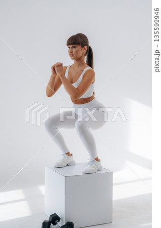 Exercise of squats on white cube. Young sportive woman is in the studio Exercise of squats on white cube. Young sportive woman is in the studio 135599946