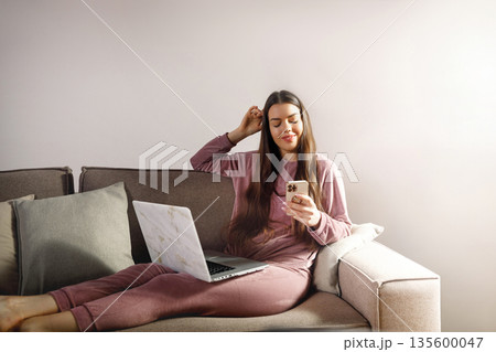 Woman sits on couch with laptop and phone, enjoying a moment of leisure at home in a comfortable setting, absorbing the online world while relaxing at ease Woman sits on couch with laptop and phone, enjoying a moment of leisure at home in a comfortable setting, absorbing the online world while relaxing at ease 135600047