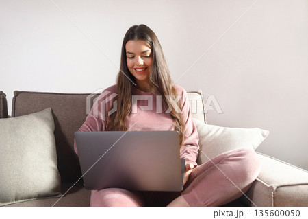 Woman sits on a couch using a laptop in a modern living room during the day while wearing comfortable clothing and smiling Woman sits on a couch using a laptop in a modern living room during the day while wearing comfortable clothing and smiling 135600050