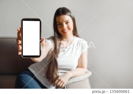 Woman sitting on couch holds a smartphone in her hand while smiling at the camera in a well-lit indoor space in the afternoon 135600061