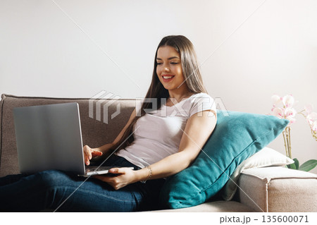 Woman sits on couch and works on laptop while smiling, enjoying a relaxing moment in a cozy living room setting with a soft pillow and flowers nearby Woman sits on couch and works on laptop while smiling, enjoying a relaxing moment in a cozy living room setting with a soft pillow and flowers nearby 135600071