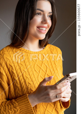 Woman in a yellow sweater smiles while looking at a smartphone in a warm indoor space during the afternoon 135600077