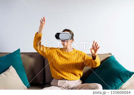 A woman is using a virtual reality headset while moving her hands to engage with the visual content. She is inside a room with light walls and a simple background. A woman is using a virtual reality headset while moving her hands to engage with the visual content. She is inside a room with light walls and a simple background. 135600084