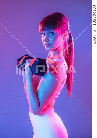 Arms exercise using dumbbell. Young sportive woman is in the studio 135600133