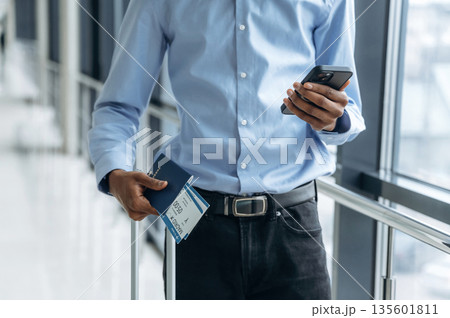 Smartphone and tickets in hands. Male tourist is in the airport corridor Smartphone and tickets in hands. Male tourist is in the airport corridor 135601811