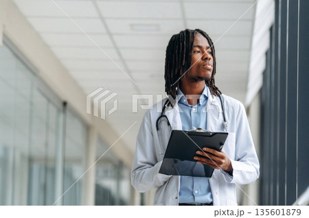 Stethoscope on neck, notepad in hands. Young male black doctor is in the corridor Stethoscope on neck, notepad in hands. Young male black doctor is in the corridor 135601879