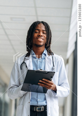 Stethoscope on neck, notepad in hands. Young male black doctor is in the corridor 135601880