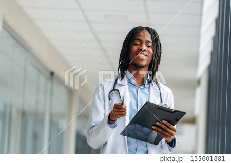Stethoscope on neck, notepad in hands. Young male black doctor is in the corridor 135601881