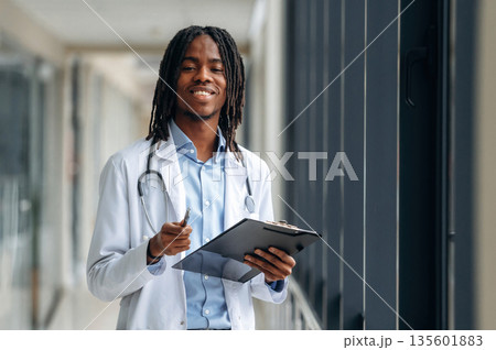 Stethoscope on neck, notepad in hands. Young male black doctor is in the corridor 135601883