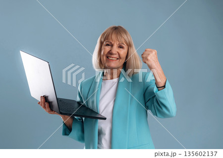 Celebrating success, holding laptop. Senior woman is against background Celebrating success, holding laptop. Senior woman is against background 135602137