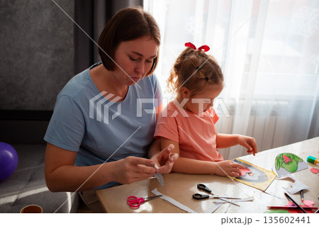 Portrait of Caucasian adult mother plays and teaches little girl how to make postcards for Mother's Day and Valentine's Day. Family sitting at the table. Preschool education	 135602441