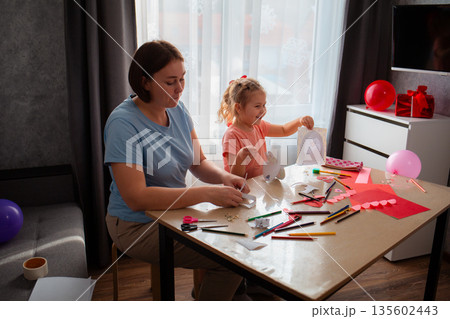 Wide shot of kindergarten teacher plays and teaches cute little happy girl how to make paper cards for Mother's Day and Valentine's Day. Woman and child sitting at the table	 135602443