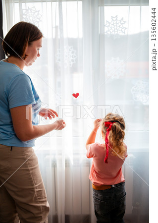 A young Caucasian mother and her daughter decorate the curtains on the window with red paper hearts. A celebration for Valentine's Day and Mother's Day. Vertical	 135602444