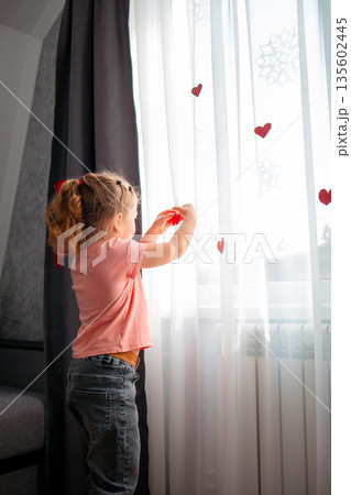 Rear view of little girl decorate the curtains on the window with red paper hearts. A celebration for Valentine's Day and Mother's Day. Vertical	 135602445